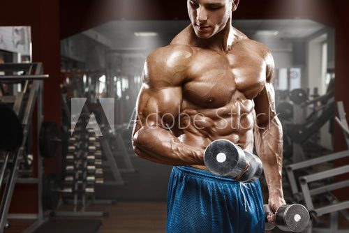 Muscular man working out in gym doing exercises with dumbbells at biceps, strong male naked torso abs